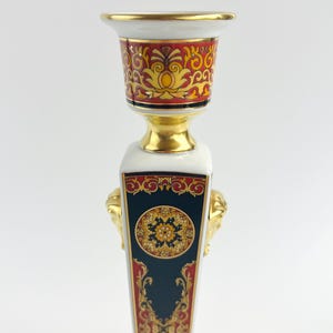 May include: A decorative ceramic candlestick with a gold rim and accents. The candlestick features a white base with a dark blue panel, gold lion head details, and red and gold ornate patterns. The top has a red and gold design.