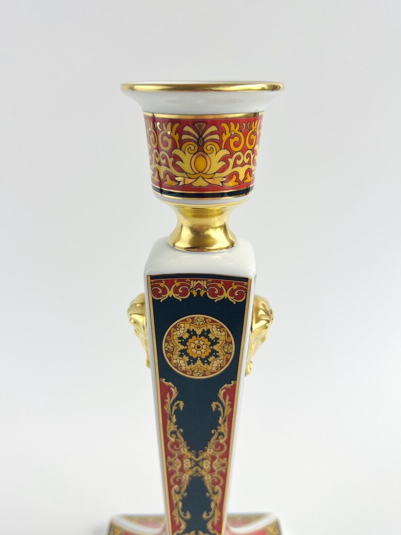 May include: A decorative ceramic candlestick with a gold rim and accents. The candlestick features a white base with a dark blue panel, gold lion head details, and red and gold ornate patterns. The top has a red and gold design.
