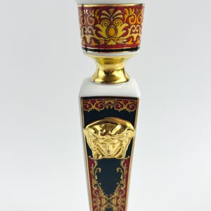 May include: A decorative candlestick with a gold, red, and black design. The candlestick features a gold Medusa head emblem and ornate patterns. The top and base have gold accents, and the overall design is symmetrical.