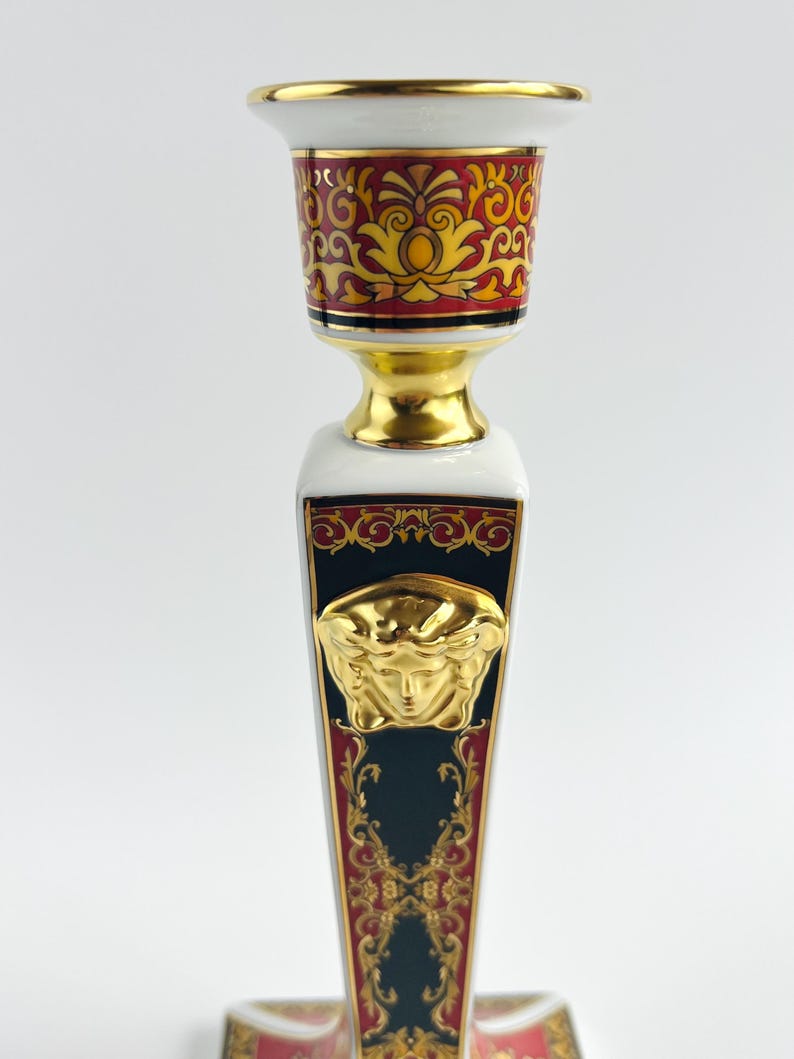 May include: A decorative candlestick with a gold, red, and black design. The candlestick features a gold Medusa head emblem and ornate patterns. The top and base have gold accents, and the overall design is symmetrical.