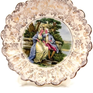 May include: A decorative porcelain plate with a scalloped edge and gold floral detailing. The center features a painted scene of a couple in period clothing. The plate is supported by a white stand.