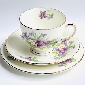 Peut inclure: Service à thé vintage composé d'une tasse, d'une soucoupe et d'une assiette. La porcelaine ivoire présente un motif floral de violettes violettes et de feuilles vertes. Des bordures dorées ornent les bords de la tasse et des assiettes.