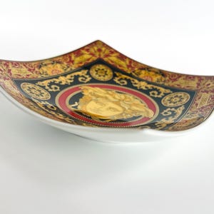 May include: A decorative, square-shaped ceramic dish with a white base and a detailed, ornate design. The rim features a black, gold, and red pattern with a central medallion. The medallion has a gold figure on a red background.