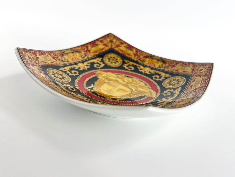 May include: A decorative, square-shaped ceramic dish with a white base and a detailed, ornate design. The rim features a black, gold, and red pattern with a central medallion. The medallion has a gold figure on a red background.