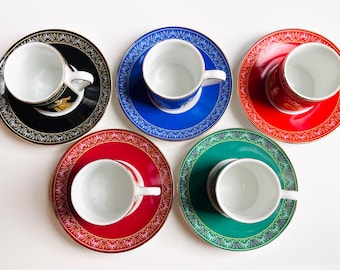 May include: Overhead shot of five teacups and saucers in a variety of colors: black, blue, red, green, and red. Each saucer has a decorative gold pattern around the rim. The teacups are white with a white handle.