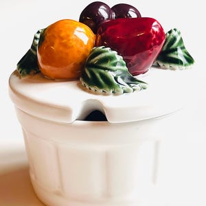May include: A white ceramic jar with a lid, decorated with ceramic fruit including an orange, a red apple, and dark purple cherries. Green ceramic leaves surround the fruit. The jar has a small opening on the lid.