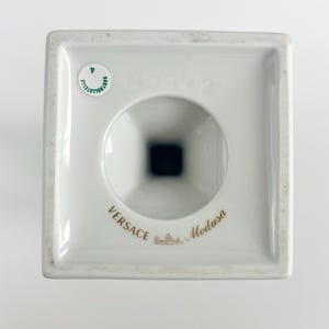 May include: A white porcelain Versace Medusa candle holder. The square base has a circular opening and a black interior. The words "Versace" and "Medusa" are printed in gold. A small sticker is visible.