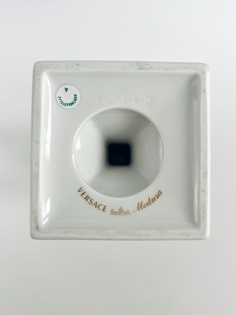 May include: A white porcelain Versace Medusa candle holder. The square base has a circular opening and a black interior. The words "Versace" and "Medusa" are printed in gold. A small sticker is visible.