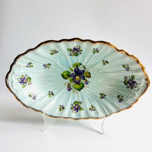 May include: Oval-shaped, light blue ceramic serving platter with a gold rim and a floral design. The platter features a central cluster of purple violets with green leaves and smaller violet clusters scattered across the surface.
