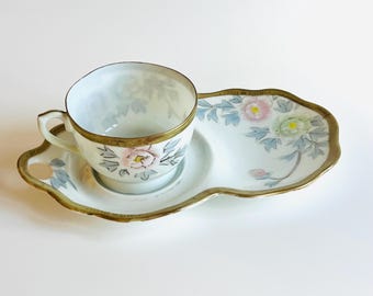 May include: A vintage teacup and saucer set. The white porcelain cup and saucer feature a floral design with pink and green flowers, gray leaves, and gold trim. The saucer is an elongated shape with a handle.