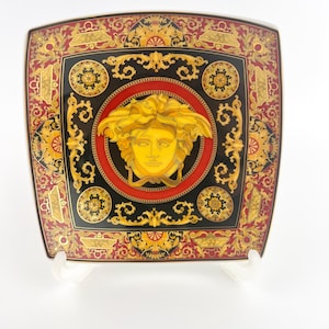May include: Square decorative plate with a black, red, and gold design. The plate features a central medallion with a gold face and intricate patterns. The plate has a square shape with rounded corners.
