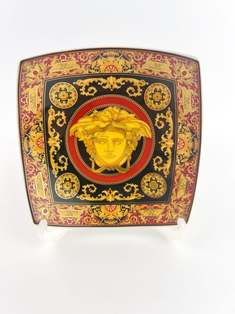 May include: Square decorative plate with a black, red, and gold design. The plate features a central medallion with a gold face and intricate patterns. The plate has a square shape with rounded corners.