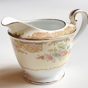 Może przedstawiać: Kremowa porcelanowa mleczarka z kwiatowym wzorem. Mleczarka posiada delikatny uchwyt i dzióbek, ze złotym wykończeniem i ozdobnym paskiem wokół krawędzi. Bukiet kolorowych kwiatów zdobi bok.