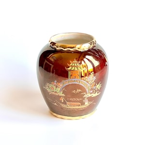 Op de afbeelding: Een decoratieve, glanzende bruine keramische vaas met een gouden rand en voet. De vaas heeft een gedetailleerde illustratie van een brug, pagode en bootscène in goud, groen en blauw. De vaas heeft een ronde vorm.