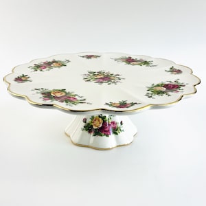 Puede incluir: Un soporte para pasteles de cerámica blanca con borde festoneado y un estampado floral con rosas rosas, amarillas y verdes. El soporte tiene un borde dorado y una base de pedestal, ideal para exhibir pasteles.