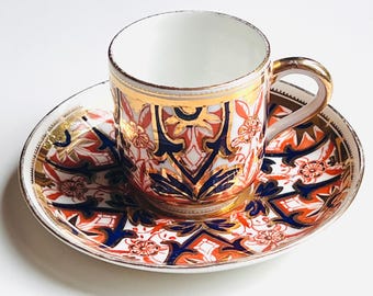 May include: A vintage teacup and saucer set. The cup and saucer feature a white background with intricate patterns in orange, navy blue, and gold. The cup has a gold handle and rim, and the saucer has a matching gold trim.