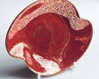 May include: A red decorative dish with a gold floral pattern and a gold rim. The dish has a unique shape with a wavy edge. The dish is displayed on a white stand.