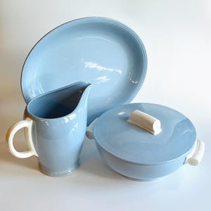 May include: A light blue ceramic serving set, including an oval platter, a covered bowl with a white handle, and a pitcher with a white handle. The set is designed for serving food and has a classic, elegant aesthetic.