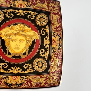May include: Square decorative plate with a black, red, and gold design. The plate features a central Medusa head surrounded by ornate patterns and a gold border. The plate has a luxurious and elegant aesthetic.