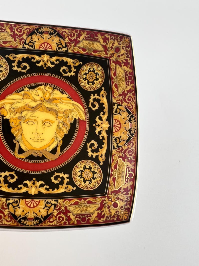 May include: Square decorative plate with a black, red, and gold design. The plate features a central Medusa head surrounded by ornate patterns and a gold border. The plate has a luxurious and elegant aesthetic.