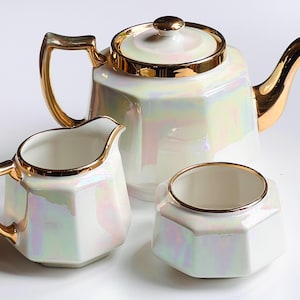 May include: A white, iridescent tea set with gold detailing. The set comprises a teapot, a creamer jug, and a sugar bowl. The teapot has a lid and a curved spout. The creamer has a handle and a pouring spout. The sugar bowl is a small, open container.