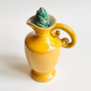 May include: A yellow ceramic jug with a handle and spout, topped with a green artichoke-shaped stopper. The jug has a glossy finish and is designed for serving drinks or as a decorative piece. The jug's height is approximately 15 cm.