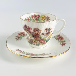 Puede incluir: Juego de taza y platillo blancos con ribete dorado. La taza y el platillo presentan un diseño floral con una ilustración de hadas. La taza tiene una textura acanalada. El juego está sobre un fondo blanco.