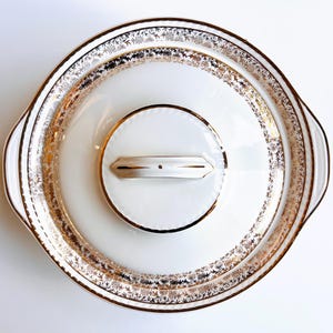 Pode incluir: Uma travessa branca com tampa, com padrões florais e geométricos dourados nas bordas. A travessa tem duas alças e uma tampa redonda pequena com uma alça dourada. Design vintage e elegante.