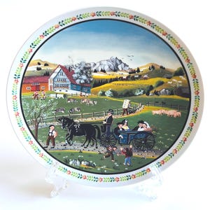 May include: A decorative plate with a pastoral scene. The plate features a colorful painting of a horse-drawn carriage, houses, and rolling hills. The rim is decorated with a floral pattern. The plate is displayed on a clear stand.