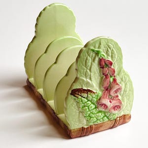 Puede incluir: Un tostador de cerámica verde claro con un diseño floral. El estante tiene cuatro ranuras y está decorado con un relieve en relieve de flores y hojas rosas y verdes. La base es marrón.
