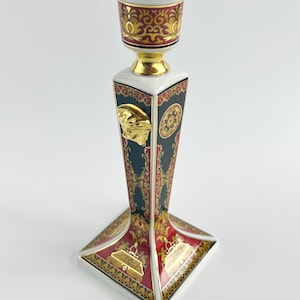 May include: A decorative ceramic candlestick with a square base and a tall, angular body. The candlestick features a gold Medusa head emblem, ornate red and gold patterns, and a white rim. The top is a circular cup with a gold rim.