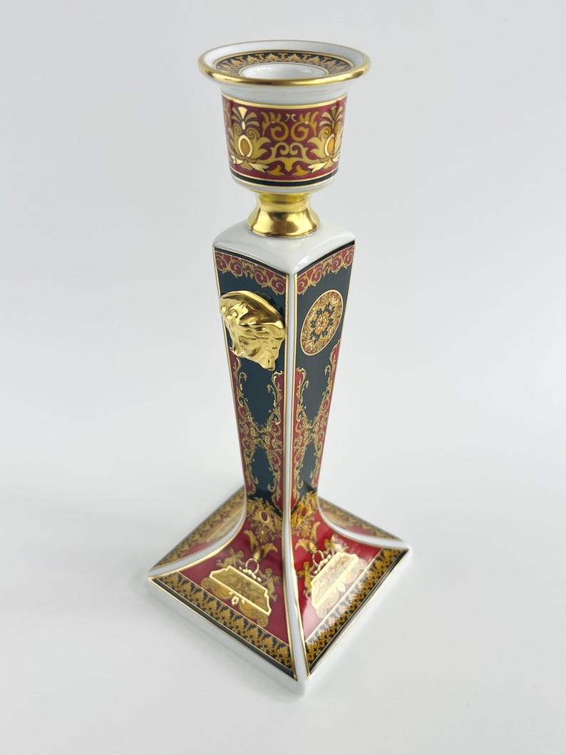 May include: A decorative ceramic candlestick with a square base and a tall, angular body. The candlestick features a gold Medusa head emblem, ornate red and gold patterns, and a white rim. The top is a circular cup with a gold rim.