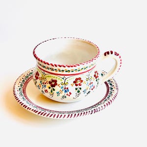Könnte beinhalten: Ein weißes Keramik-Tee-Tassen- und Untertassen-Set. Die Tasse und die Untertasse sind mit farbenfrohen Blumenmustern in Rot, Grün, Blau und Gelb verziert. Der Rand der Tasse und der Untertasse hat eine rot-grüne Verzierung.