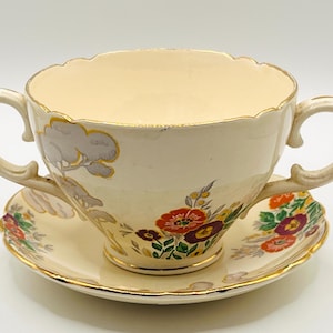 Puede incluir: Taza de té color crema con dos asas y platillo a juego. El juego presenta diseños florales y de árboles en rojo, morado y dorado. Los bordes son festoneados y ribeteados con oro.