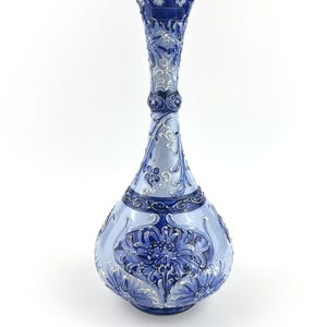 May include: A tall, slender ceramic vase featuring a bulbous base and a narrow neck. The surface is decorated with intricate, raised relief patterns of blue and white floral motifs, with deep blue accents on the rim and base sections.