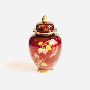 Puede incluir: Un jarrón de cerámica granate con tapa, con un remate y base dorados. El jarrón está adornado con un diseño floral en amarillo, rosa y blanco, con detalles dorados. Es un objeto decorativo.