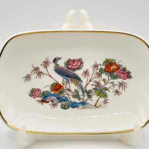 Puede incluir: Un plato rectangular de cerámica blanca con borde dorado. El plato presenta una colorida ilustración de un pájaro posado en una rama con flores. El pájaro tiene detalles azules, rojos y amarillos. Las flores son rosas y naranjas.