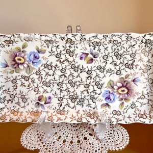 May include: Rectangular ceramic serving tray with a floral pattern. The tray features a white background with a repeating pattern of small black flowers and larger purple and blue floral arrangements. The edges are scalloped.