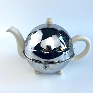 May include: A spherical, chrome teapot with a white spout, handle, and lid knob. The teapot sits on three small, white feet. The chrome surface reflects the surrounding environment.