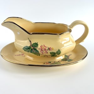 May include: A cream-coloured gravy boat and saucer set. The boat has a curved spout and handle, with a floral design of green leaves and pink flowers. Gold trim accents the edges of both pieces, creating an elegant look.