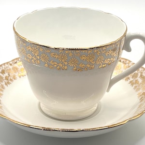 Puede incluir: Una taza y un platillo blancos con detalles florales dorados. La taza tiene un asa curva y un borde dorado. El platillo tiene un borde festoneado y un patrón floral dorado a juego. Un juego de té clásico.