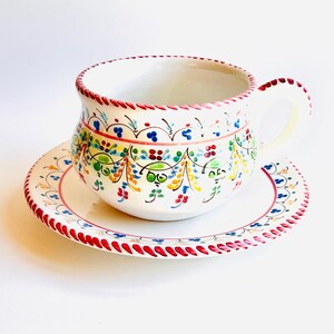 Könnte beinhalten: Ein weißes Keramik-Tee-Tassen- und Untertassen-Set. Die Tasse und die Untertasse sind mit bunten Blumenmustern in Blau, Grün, Gelb und Rot verziert. Der Rand der Tasse und der Untertasse ist mit einem roten Punktmuster verziert.