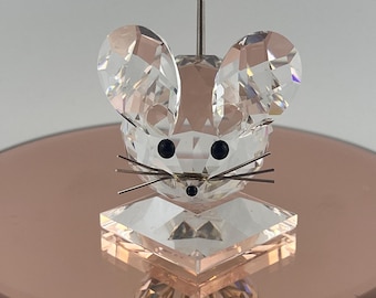 May include: A clear faceted crystal figurine of a mouse head with two large rounded ears, two black circular eyes, a small black circular nose, and three thin metallic whiskers on each side, all mounted on a square clear crystal base.
