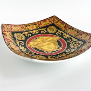 May include: Square decorative dish with a white base and a black, red, and gold ornate design. The center features a gold Medusa head on a red background. The dish has a unique, curved shape.