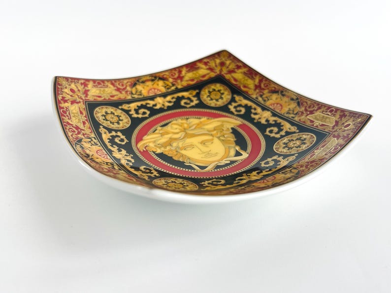 May include: Square decorative dish with a white base and a black, red, and gold ornate design. The center features a gold Medusa head on a red background. The dish has a unique, curved shape.