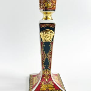 May include: A decorative ceramic candlestick holder with a gold Medusa head emblem. The candlestick features a white, red, and gold color scheme with ornate detailing. The base is square, and the top is designed to hold a single candle.