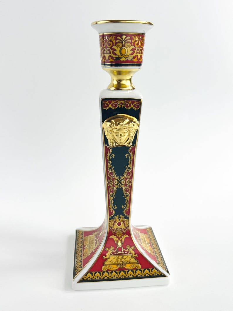 May include: A decorative ceramic candlestick holder with a gold Medusa head emblem. The candlestick features a white, red, and gold color scheme with ornate detailing. The base is square, and the top is designed to hold a single candle.