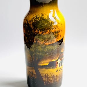 May include: A ceramic vase with a hand-painted rural landscape scene. The vase features a sunset with a farmhouse, trees, and chickens. The colors include shades of yellow, orange, brown, and green. The vase has a dark brown rim and base.