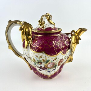 Pode incluir: Uma bule de porcelana decorativo com um design bordô e dourado. O bule apresenta um padrão floral na metade inferior e detalhes em folha de ouro na alça e no bico. A tampa é bordô com um botão dourado.