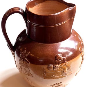 Pode incluir: Um jarro de cerâmica castanho com uma pega e uma abertura larga. O jarro apresenta cenas em relevo de um moinho de vento, pessoas e um barril. O jarro tem um acabamento brilhante e um esquema de cores de dois tons.
